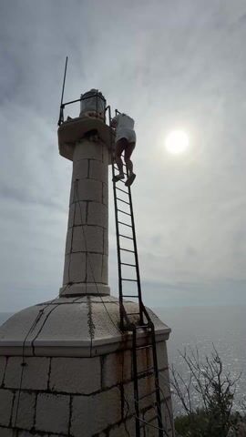 A low-angle shot captures a person climbing a metal ladder attached to a stone lighthouse. The person, dressed in a white top and shorts, is nearly at the top of the ladder, reaching for the lighthouse's lantern room. The lighthouse stands against a backdrop of a cloudy sky and the ocean. The sun is visible behind the clouds, creating a bright glow.