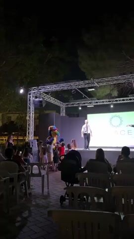 A group of children are gathered around a costumed character, possibly a mascot, on an outdoor stage at night. The character, which appears to be a bird with a colorful beak and hat, is interacting with the children. The stage is lit by spotlights, and a screen behind the character displays a logo. An adult, dressed in a white shirt and tie, stands on the stage to the right of the mascot, observing the interaction. The audience, including adults and children in strollers, is seated in chairs facing the stage. Trees and buildings are visible in the background, illuminated by artificial lighting.