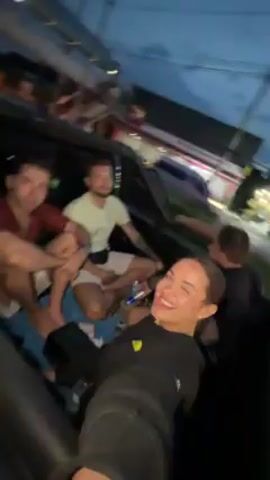 A group of four people ride a water ride at night. The camera is hand-held and pointed back at the group by the woman in the front. She smiles and makes hand gestures towards the camera. The two men in the middle of the boat sit next to each other and smile. The fourth person is partially visible behind the woman. The surrounding area is dark.