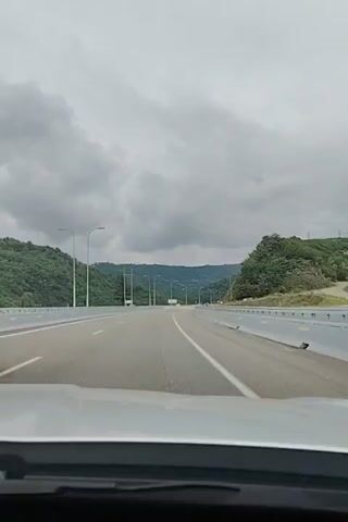 A dashcam video captures a highway scene on a cloudy day. The camera is inside a vehicle, looking forward. A red car is visible in the left lane of the highway, moving away from the camera. The highway has multiple lanes and is surrounded by lush green mountains. The sky is overcast with heavy gray clouds. Streetlights are visible on poles along the side of the highway. The road appears to be a modern highway with concrete barriers and clear lane markings.