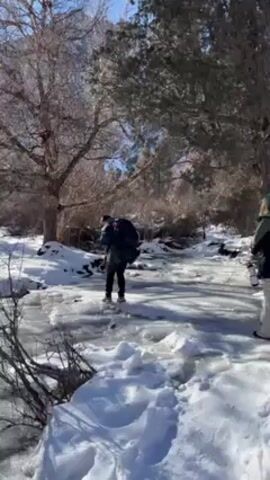 A person wearing a dark jacket and backpack walks across a partially frozen stream or river in a snowy landscape. The ice appears thin and has open water areas. The person is walking carefully across the ice, with another person standing nearby on the snowy bank. Trees surround the area, and the sky is visible in the background. The person walking on the ice slips slightly and uses their arms for balance but continues to walk forward. Someone can be heard speaking in the background.