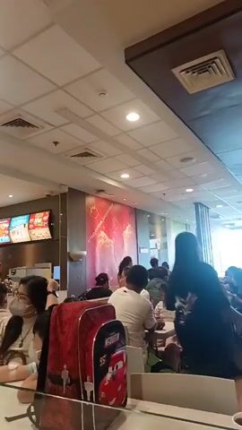 The video shows the interior of a busy fast-food restaurant. In the foreground, a young boy wearing a turquoise shirt is seated at a table, looking off to the right. Behind him, a person in a white t-shirt leans over a table, seemingly looking at a phone. Further back, other patrons are visible seated at tables and at the counter. Menu boards with colorful images of food are displayed above the counter. Red stanchions with numbers are placed on tables, likely indicating seating assignments or order numbers. The lighting appears to be from overhead fluorescent fixtures. The overall atmosphere suggests a public dining establishment with ongoing customer activity.