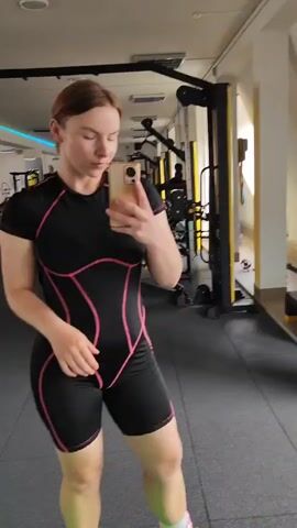 The media file shows a young woman with auburn hair standing in a gym. She is wearing a black and pink athletic outfit that appears to be a one-piece suit with short sleeves and shorts. She is holding a smartphone in her right hand and taking a selfie, looking at the phone screen. The woman has a neutral expression.

The setting is a gym with various exercise equipment visible in the background. There is a weight machine with black and yellow accents directly behind her, and other gym equipment further in the background, including what looks like a cable crossover machine and some treadmills. The floor is dark grey. The lighting in the gym suggests it might be daytime, though it's difficult to determine the exact time of day. The overall atmosphere is that of a fitness facility. There is no visible text in the media file.