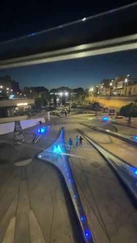 This media file captures an evening scene in Nicosia, Cyprus, showcasing a modern public plaza illuminated by blue lights. The time of day is clearly dusk or early night, indicated by the dark sky and artificial lighting. The weather appears clear.

The primary subjects are a few individuals walking through the plaza. They are casually dressed, suggesting a relaxed atmosphere. The plaza itself is the most prominent object, featuring a striking design with geometric shapes and integrated water features that are lit with vibrant blue LEDs. These water features appear to be fountains, adding a dynamic element to the scene.

In the background, buildings line a street, hinting at the urban setting. Some of these buildings have lights on, suggesting that businesses or residences are active. Streetlights are also visible, contributing to the overall illumination of the area. Trees are scattered throughout the plaza and along the street, adding greenery to the urban landscape.

The activity taking place is simply people enjoying the plaza in the evening. The overall emotion conveyed is one of tranquility and modern urban life. The blue lighting creates a contemporary and somewhat futuristic aesthetic.

Visible text includes "CAFFE NERO" on a building in the background, identifying a coffee shop. The design of the plaza itself, with its sharp angles and sleek lines, is a notable detail that suggests a recent or contemporary urban development project. The overall impression is of a well-maintained public space designed for evening leisure and enjoyment.