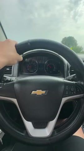 The video shows a first-person perspective of someone driving a Chevrolet. The driver's left hand is visible on the steering wheel, which has a black cover. The dashboard displays the speedometer and tachometer, both showing readings below 40. The car appears to be on a highway with other vehicles visible in the distance. The sky is cloudy. The scene is well-lit, suggesting daytime.