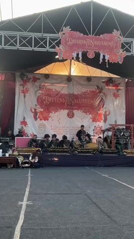A group of musicians performs on an outdoor stage under a canopy. The stage is decorated with a large banner that reads "DHARMA NAWASEN" and features floral motifs. Several musicians are seated on the stage, playing traditional Indonesian instruments, including what appear to be gamelan instruments. One musician stands and plays a wind instrument, possibly a saxophone. The musicians are dressed casually. In the foreground, there is a paved area with white lines. The background shows a brightly lit sky, suggesting it is daytime.
