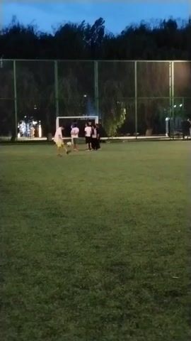 A group of five young people are playing soccer on a green field. They are standing near a soccer goal. The sky is dark, suggesting it is evening. The field is lit by artificial lights.