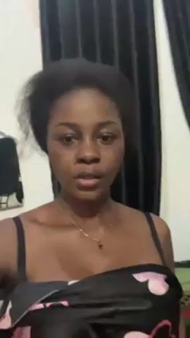 The video shows a woman indoors, standing in front of a white wall and black curtains. She is wearing a dark top with a pink floral pattern and a thin gold necklace. Her hair is dark and somewhat disheveled. She holds up a bright green plastic bag to the camera, then lowers it. She speaks directly to the camera. A clothing rack is visible in the background to the left.