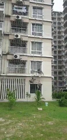 The video shows a tall apartment building with a person on a makeshift scaffolding, cleaning the exterior of a window. The person is wearing a dark outfit and is connected to the scaffolding with a rope. Buckets hang from the scaffolding, presumably containing cleaning supplies. Another person is also visible on a lower section of the scaffolding, possibly assisting or waiting. The building has multiple windows, air conditioning units, and balconies. The sky is overcast.