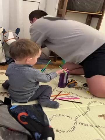 A person and toddler are on a patterned floor mat in a room. The toddler is in a seated position wearing a gray long-sleeved shirt, gray pants, and gray socks. The person is kneeling near the toddler, wearing a light gray t-shirt and black shorts. Both are facing towards a purple bottle with colorful straws near it. The toddler places a straw into the bottle, then picks up a small dark-colored toy. A toy donkey is positioned behind the toddler. The room also contains toy vehicles, pipe cleaners, and a playpen. There is a wooden shelf and a white wall visible. The person is talking to the toddler.