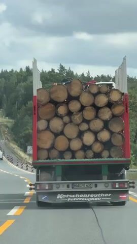 The video shows a truck carrying a load of cut logs on a highway. The truck is in the right lane, and other vehicles are visible in the lanes to the left. The truck has a German license plate that reads "MAB FB 631" and displays the company name "Kotschenreuther Fahrzeugbau" on the back. The logs are of varying sizes and are neatly stacked in the trailer. The environment appears to be a wooded area with trees lining the highway, and the sky is cloudy. The camera is positioned behind the truck, suggesting it is from another vehicle on the road. The video appears to be taken during the daytime.