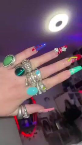 A hand wearing numerous rings and a charm bracelet is shown in a short video. The nails are long and decorated with colorful designs, including dice, a martini glass, and the word "lucky". The background is a room with a bright light and a purple and red glow. There is music playing.