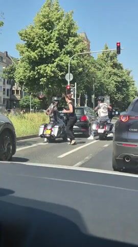 A woman in black stands to the left of a motorcycle. She is facing away from the camera and appears to be talking to the rider. The motorcycle rider is wearing a helmet and a black vest. Another motorcycle with a rider wearing a similar outfit is to the right, further up the road. Both motorcycles are facing forward in what appears to be a traffic lane. A black car is stopped in front of the first motorcycle. The traffic light is red. Trees and buildings are visible in the background. The scene is taking place outdoors on a sunny day.