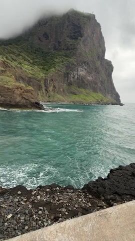 The video captures a view of a coastal scene on an overcast day. Dominating the background is a massive, sheer cliff face, partially shrouded in fog or low-lying clouds. The cliff is rugged, with patches of green vegetation clinging to its slopes. The sea in the foreground is a vivid turquoise color, with small whitecaps forming as the water laps against the rocky shoreline. The foreground is composed of black volcanic rock and pebbles, with a concrete edge visible, suggesting a man-made structure like a pathway or a wall. There are no people or animals visible in the video. The scene is serene and emphasizes the rugged natural beauty of the coastline.