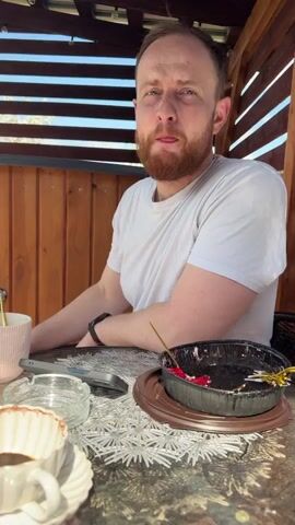 A man sits at an outdoor table with a half-eaten cake, a coffee cup, an ashtray, and a phone. He wears a white t-shirt and has a beard. He looks at the camera and then at the cake. He pokes the cake with a small gold spoon. The table is covered with a silver textured placemat. The man is sitting under a wooden structure with horizontal slats. The light is bright and sunny.