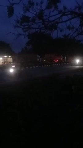 The video shows a dark, outdoor scene at night, possibly dusk or early morning. Silhouettes of trees are visible against a dark blue sky. In the background, there are lights from what appear to be buildings or stalls, and several trucks are parked or driving. One truck's headlights are on and illuminate the road. The ground in the foreground is dark and indistinct.