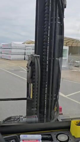 The video shows a first-person perspective from inside a forklift or similar heavy machinery, moving through an outdoor industrial area. The view is framed by the forklift's mast, which features chains and hydraulic lines. In the foreground, there is a yellow holder containing pens. The forklift is moving forward, and the environment consists of a paved area with yellow lines, stacks of white-wrapped material, a large industrial building on the left, and a covered structure with white walls and green windows in the distance. The sky is overcast. The forklift appears to be in motion, navigating the paved area between the stacks of materials.
