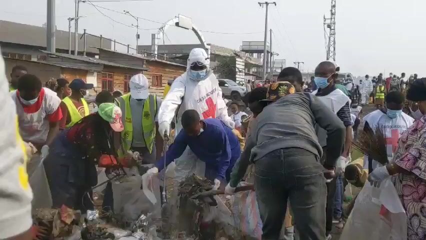 A group of people are cleaning up a large pile of garbage in an urban environment. Many of the individuals are wearing face masks and gloves. One person is wearing a white hazmat suit with a red cross on the front. Several others have vests with "Croix-Rouge" (Red Cross) printed on them. The garbage pile appears to consist of plastic bags, bottles, and other discarded items. Some people are using shovels to gather the debris into bags. In the background, there are buildings, a utility pole, and a tower. The overall impression is that this is a coordinated effort to clean up a heavily littered area.