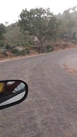 The video shows a view from a moving vehicle, possibly a car, traveling on a paved road through a mountainous or hilly area. The sky is overcast and there appears to be some fog or haze in the distance. The roadside is lined with dry, brown grass and sparse trees. Concrete barriers run along sections of the road. Some graffiti is visible on the concrete barrier in the middle of the video. The terrain of the mountains is dry and rocky. The side mirror of the car is visible in the lower left corner of the screen in a few shots.