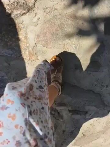 The video opens with a close-up shot of a person's legs and feet walking on a cobblestone path. The person is wearing a floral-patterned dress. The camera then pans up to reveal the path winding uphill, lined with stone walls and lush greenery. In the background, a building with a wooden roof and windows is visible, surrounded by trees and flowers. The sky is a clear blue. The video appears to be shot on a sunny day, capturing the charm of a quaint, possibly Mediterranean, village.