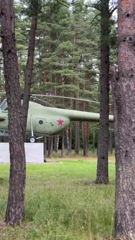 A green Soviet-era helicopter, identified by a red star insignia and the number "01" on its side, is displayed on a concrete pedestal in a grassy area surrounded by trees. The helicopter appears to be stationary, with its rotors still. A white informational plaque with images and text in Russian is attached to the front of the pedestal. The scene is filmed from a moving perspective, indicating the viewer is likely walking or being driven past the display. The weather appears overcast, and the foliage suggests a temperate climate.