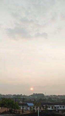 A vertical video begins with a close-up of a rooftop, then pans up to reveal a hazy sky with faint clouds. The sun is visible as a small, bright orb in the center of the frame, low on the horizon. Below the sky, a silhouette of a town with buildings and trees can be seen. The video appears to capture either sunrise or sunset.
