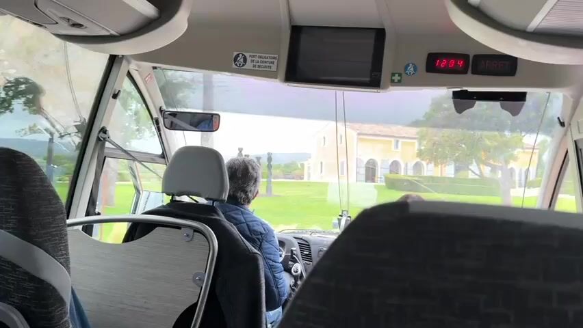 The view from inside a passenger bus or van, traveling towards an orange-toned, two-story building with arched entrances. Two passengers are visible in the front seats, their backs to the camera. The driver has short, gray hair and wears a blue quilted jacket. The building is set within a large grassy area, surrounded by trees and bushes. Beyond the building and grounds, a rural landscape with vineyards and trees extends into the distance. The bus is equipped with an overhead screen and digital clock displaying "12:04". A sign is visible stating "PORT OBLIGATOIRE DE LA CEINTURE DE SECURITE". The weather appears overcast or rainy, as evidenced by moisture on the windshield.