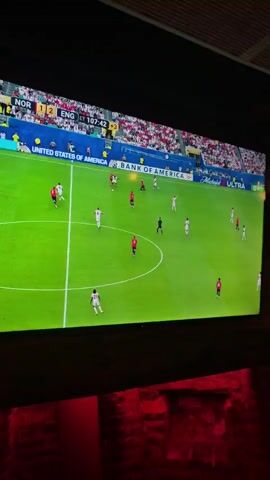 The video shows a bar scene with a large screen displaying a soccer match with the score NOR 1 - 2 ENG. There are people in the foreground watching the match. The bar has a rustic wooden interior with exposed beams and pendant lights. The background is dimly lit with purple and pink neon lights and a checkered floor. People are seen standing and talking in the bar.