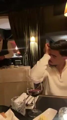 The video depicts a well-dressed waiter preparing something on a white-clothed table in a dimly lit restaurant.  The waiter, with graying hair and wearing a vest over a light-colored shirt and tie, is focused on the task at hand. Behind him, through a curtained archway, a bar with bottles is visible.  In the foreground, a man in a light-colored sweater sits at another table with a glass of red wine, napkins, and plates. The camera is at eye level, suggesting the video was taken by someone seated at the man's table.