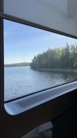 The video shows a view from inside a moving vehicle, likely a train, looking out a window. The window has a pull-down shade at the top and a speaker or vent below the glass. Outside the window, a body of water is visible on the left, with a lush green shoreline lined with trees. The trees are a mix of deciduous and coniferous, and the sun is shining, casting dappled light on the foliage. As the vehicle moves, the landscape changes. The water recedes, and the shoreline becomes steeper and rocky. There are some man-made structures visible, including what appears to be a fence and some electrical equipment. The overall impression is of a scenic journey through a natural landscape.