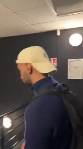 The video starts in a stairwell with dark walls, a black metal railing, and fluorescent lighting. There are two fire extinguishers on the wall along with a building layout map and other safety signs. A person wearing a white baseball cap and carrying a black backpack walks past the camera. They are walking in front of a large black backdrop with yellow illustrations of lightning bolts, hearts, tennis balls, and tennis rackets. The word “PADEL” is in white letters at the bottom.