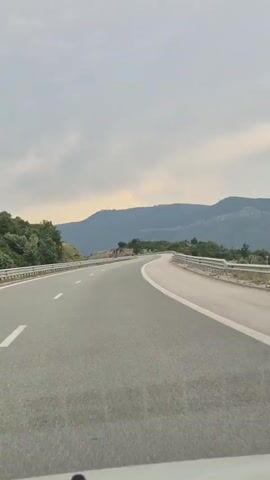 This video shows a view from a vehicle driving on a highway. The highway curves to the right, with a metal guardrail on either side. Beyond the guardrail on the left, there are trees and a rocky incline. On the right, the guardrail runs alongside a steep drop-off with a wide, hazy expanse visible in the distance. In the background, there are rolling hills and mountains.

The sky is overcast with a muted gray color, but there are hints of pale yellow and orange light, suggesting it might be late afternoon or early evening as the sun tries to break through the clouds. The overall atmosphere is calm and somewhat subdued. There is no text visible in the video. The setting appears to be a rural or mountainous area, consistent with the context of Glozhene, Bulgaria. The activity is simply driving along the highway, with the camera capturing the passing scenery.