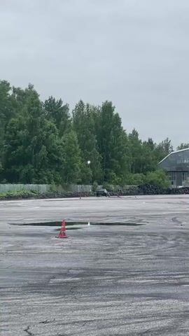 A black SUV is drifting in circles on a wet, paved surface. In the background, there is a large, metal building and a dense forest. The sky is overcast. Several orange traffic cones are scattered on the ground. The SUV appears to be the only vehicle actively drifting. The car is leaving tire marks on the asphalt. The overall scene suggests a drift racing or practice session.