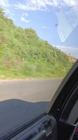 A video taken from inside a car shows a view out the passenger window. The car is moving along a paved road. To the left of the road, there is a grassy hillside covered in green vegetation, with some patches of brown earth visible. The sky above is blue with scattered white clouds. The interior of the car is visible in the foreground, including the door frame and part of the dashboard. The sun appears to be shining, casting shadows on the road.