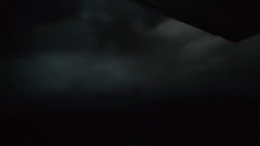 This video begins in near complete darkness, with a faint, blurry object visible in the center of the frame around the 0:00 to 0:03 mark. As the video progresses to 0:04, the scene brightens slightly to reveal dark, stormy clouds overhead. The lower portion of the frame remains dark, obscuring any ground-level details.