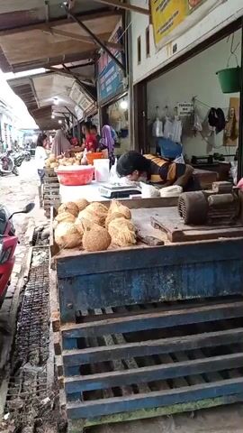 A person in a yellow and black striped shirt is weighing items on a scale at an outdoor market stall. The stall has a display of coconuts in the foreground. Other people are visible in the background, as well as motorcycles parked along a walkway. The scene appears to be a bustling marketplace with goods displayed for sale.