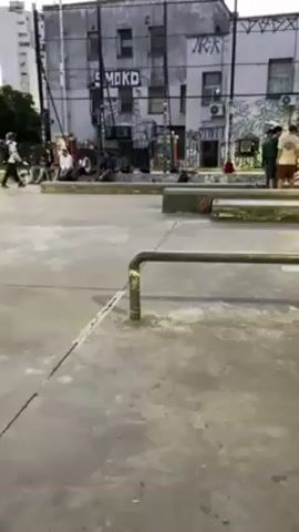 The video shows a person skateboarding on a metal rail in an urban environment. The skateboarder, wearing a green shirt and jeans, approaches the rail, ollies, slides along the rail, and rides away. The location appears to be a skate park or plaza, with concrete surfaces, other rails and platforms, and graffiti-covered buildings in the background. Other people are standing around, some observing the skateboarder. The lighting suggests it is either dusk or dawn. There are tall buildings surrounding the area.