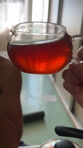 A person's hand holds a clear glass teapot filled with a dark red liquid. The teapot has a handle and a spout. The person's thumb and index finger are gripping the handle. A person's bare shoulder and upper back are visible on the left side of the frame, out of focus. The background appears to be an indoor setting with a green surface in the foreground and a window or light-colored wall in the background. The lighting suggests daylight.