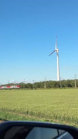 A long train with red and gray carriages passes by a field of green crops. A large white wind turbine stands tall in the background under a clear blue sky. The video appears to be filmed from a car, with the side mirror visible in the foreground.