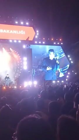 A crowd watches a male singer perform on a large stage. The stage features a large screen displaying the singer's image and Turkish text, likely related to the Ministry of Culture and Tourism. There are bright spotlights and a complex geometric light display behind the singer and his band. The crowd is densely packed, and the singer appears to be energetically performing.