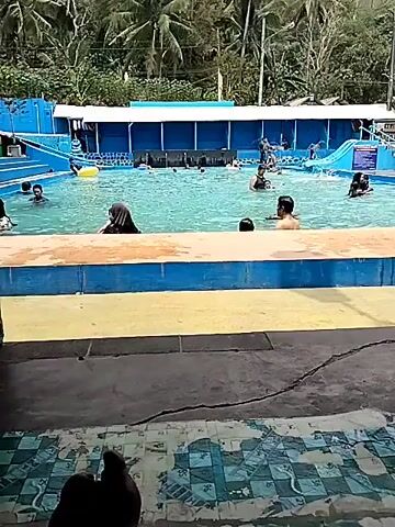 The video shows a swimming pool area on a sunny day. Several people are swimming in the bright blue water, some with inner tubes. A blue and white slide is visible in the background. On the side of the pool, there are blue seating areas with a light-colored tile floor. Some people are sitting in these areas, watching others swim. In the foreground, there is a cracked concrete area and a blue tiled floor. Trees and buildings can be seen in the background. The overall atmosphere appears to be recreational and relaxed.