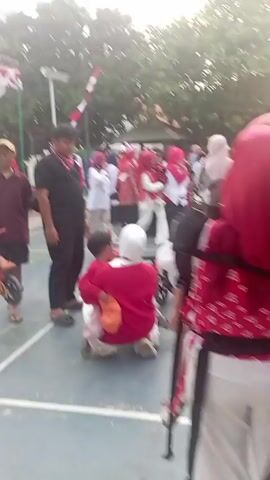 The video depicts an outdoor gathering of people, likely a community event or celebration, with a focus on children and families. The scene is a paved area, possibly a sports court, with trees in the background. Red and white decorations, including flags and banners, are prominently displayed, suggesting a national holiday or a themed event.

In the foreground, a woman wearing a red headscarf and red top is kneeling, holding a young child who is sitting on her lap. The child is wearing a pink shirt. Another child, a boy, is sitting on a small bicycle decorated with colorful streamers. The woman appears to be interacting with the child on the bicycle.

To the left of this central group, a man wearing a black t-shirt and black pants stands with his arms crossed, observing the scene. He has a red bandana around his neck. Further left, a person in a red shirt and a cap is visible.

In the background, there is a stage area decorated with red and white fabric. Several people are gathered around the stage, some wearing red and white attire. One person on the stage is holding a large flag. A stroller is also visible near the stage.

The overall atmosphere seems festive and familial, with a mix of adults and children participating in the event. The lighting suggests it is daytime, possibly overcast.