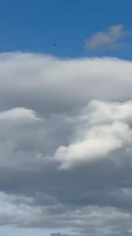 The video shows a vertical view of a blue sky with scattered white and gray clouds. Two black birds are visible flying in the distance against the blue portions of the sky, above the clouds. The clouds vary in shape and density, with some appearing brighter white and others darker gray. The sky is mostly visible above the cloud cover.