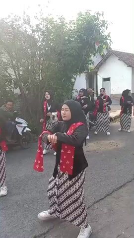A group of women in traditional Indonesian attire, including headscarves and batik skirts, are performing a dance outdoors on a paved street. They are wearing black tops with red sashes or scarves draped over their shoulders. The woman in the foreground is facing away from the camera and moving her arms and body rhythmically. In the background, other dancers are visible, as well as a man sitting on a motorcycle and a few buildings. The environment appears to be a village or rural area, with trees and vegetation present. The sun is shining, casting a bright light on the scene.