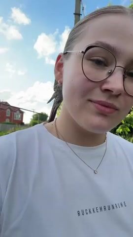 A young woman with glasses and her hair in a bun is outside in front of green foliage and a house. She is wearing a white t-shirt with text on it. She adjusts her hair and smiles at the camera.