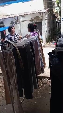 Two women are interacting in front of racks of clothing that appear to be for sale outdoors. The woman on the left, wearing a dark patterned top, is speaking to the woman on the right, who is wearing a purple top and has her back to the camera. Behind them, a man wearing a cap and a light-colored shirt is sitting on a motorcycle with a sidecar. The environment appears to be an alleyway or a market stall with dirt ground and various structures in the background. The clothing is hung on hangers and displayed on racks, with some items visible in the foreground and background.