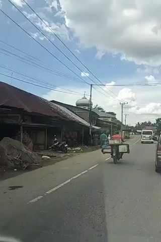 The video shows a street scene in what appears to be a developing country. A motorcycle with a sidecar, carrying a person and several containers, drives down the right side of the road. A few other motorcycles and a van are also visible on the road. On the left side of the road, there are several single-story buildings with corrugated metal roofs, some of which appear to be shops or market stalls. A mosque with a dome is visible in the background. The sky is partly cloudy. The overall impression is one of a bustling, everyday scene in a community.
