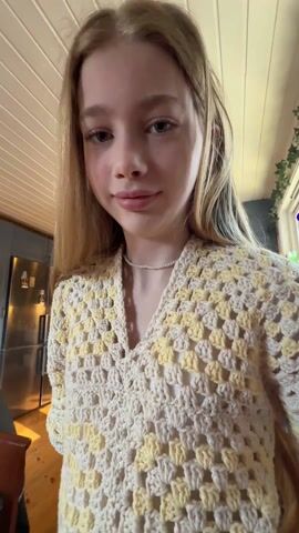 A young girl with long blonde hair is standing in a kitchen, wearing a crocheted yellow and white top and a pearl necklace. She smiles at the camera as the person filming compliments her top and mentions that she made it herself. The kitchen has a silver refrigerator, wood cabinets, and plants in the background.