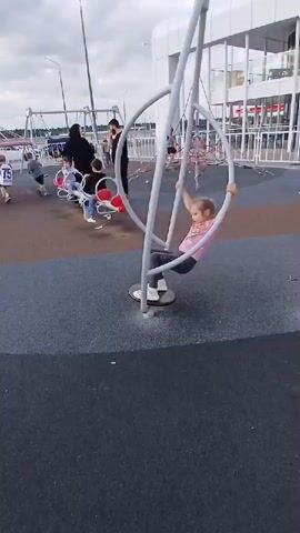 A young girl with blonde pigtails is on a playground, on a circular spinning device. She is wearing a pink shirt and grey pants. She is holding onto the metal structure of the device, her feet on the platform. In the background, there are other children on a seesaw-like swing, with adults supervising. The playground has a soft, multi-colored surface. There is a white building with large windows in the background, and a cloudy sky. The girl is actively spinning and moving on the device.