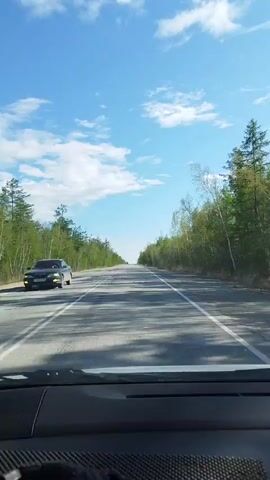 The video shows a car driving down a straight, paved road. The road is flanked by trees on both sides. The sky is blue with scattered white clouds. Another car is visible in the distance, driving in the same direction. The video is shot from the driver's perspective.